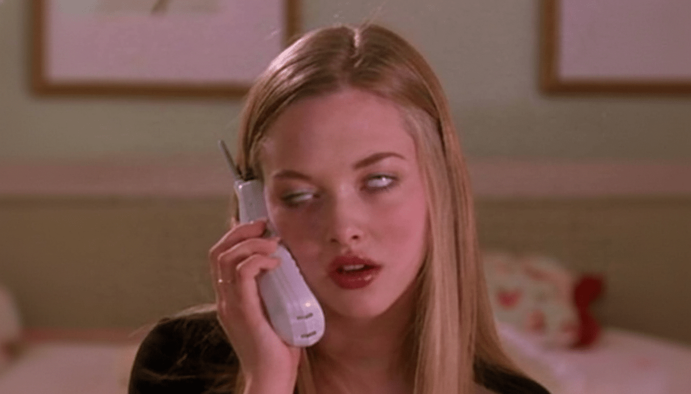 A young woman with blonde hair holds a white flip phone to her ear , rolling her eyes upwards with an exasper ated expression. The background is a soft pink room.