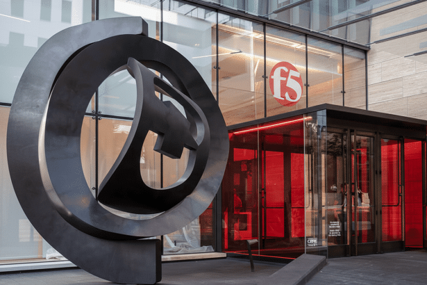 The F5 logo sculpture stands prominently in front of the modern F5 office building entrance , which features red-lit glass doors.