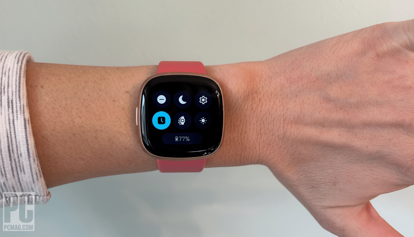 A hand wearing a pink- stra pped smartwatch with a dark screen displaying control icons and 77% battery life.