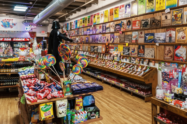 An interior view of a vibrant candy store with a wide variety of sweets , drinks , and novelty items displayed on wooden shelves and tables . A life -sized Darth Vader statue stands prominently near large l ollipops, and the walls are adorned with an array of retro posters and advertisements.