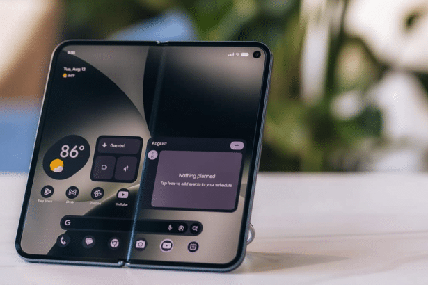 A modern foldable smartphone with a dark interface displaying widgets for weather, apps , and a calendar with Nothing planned for August. The phone is pro pped up on a surface with a blurred green and white background.