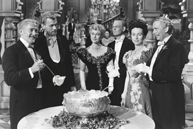 A black and white image of six formally dressed people standing around a large ornate punch bowl, appearing to be at a fancy event or party .