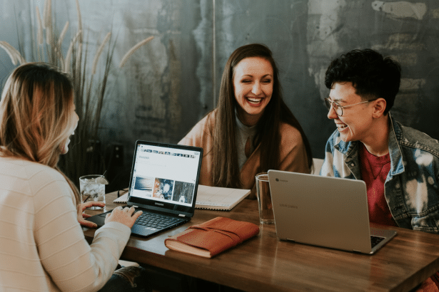 Three people laughing and working on laptops at a table in a cafe setting , resized to a 1 6:9 aspect ratio.