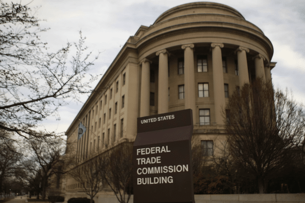 A 16:9 aspect ratio image of the Federal Trade Commission Building with its sign in the foreground and bare trees under a cloudy sky.