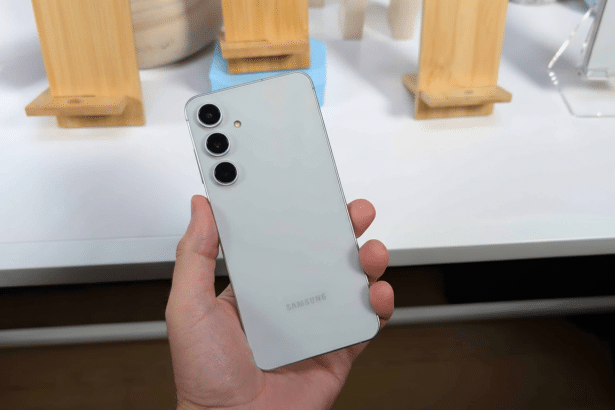 A hand holds a light gray Samsung smartphone with three cameras on the back, presented in a 16: 9 aspect ratio, maintaining the original background.