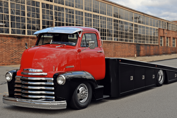 A vintage red and black Chevrolet truck with a flatbed trailer, parked in front of a brick building with many windows .