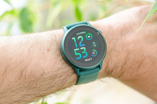 A professional shot of a Garmin smartwatch with a teal band on a person 's wrist, showing the time 12:5 3, the date Wednesday 9 , and activity icons , set against a blurred background of green leaves.