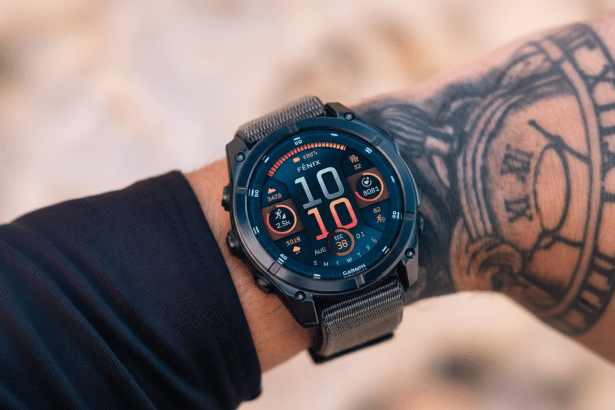 A close -up shot of a Garmin Fenix smartwatch on a person' s tattooed wrist, displaying time , activity metrics, and other data on its colorful interface , with a blurred outdoor background. Filename : garmin fenixsmart watchwrist. png