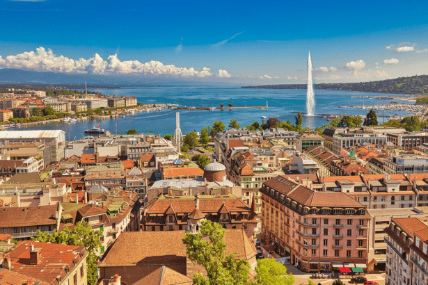An aerial view of Geneva, Switzerland , showing the city ' s buildings , Lake Geneva, and the Jet d 'Eau fountain under a clear blue sky. Filename : geneva cityscape lake jetde au .png