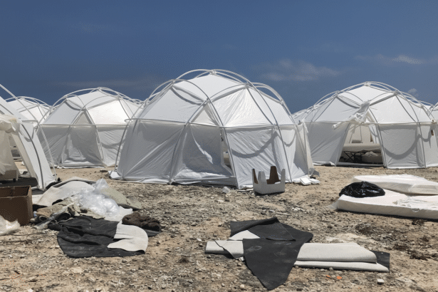 A group of white geodesic dome tents set up on a rocky, uneven terrain under a clear blue sky, with some debris scattered in the foreground.