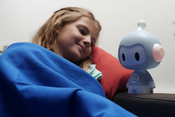 A young girl, covered in a blue blanket and resting on a red pillow, smiles gently while looking at a small, light blue robot with pink ears and a single antenna , sitting on a dark couch beside her. Filename : girl androbot oncouch .png