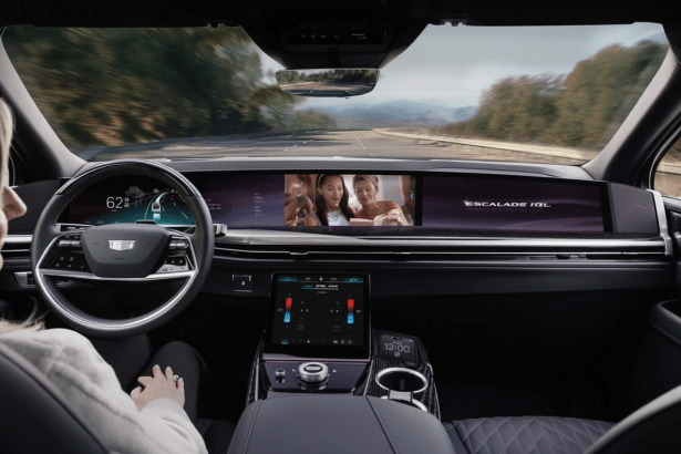 Interior view of a Cadillac Escal ade IQ from the driver's perspective, showcasing the large dashboard screen and a woman ' s hands on the steering wheel.