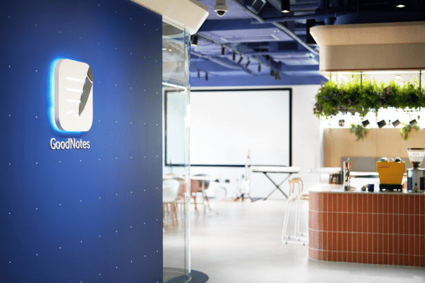 The Good Notes logo displayed prominently on a blue wall in a modern office or co -working space, with a cafe counter and meeting area visible in the background . The image has been resized to a 16: 9 aspect ratio.