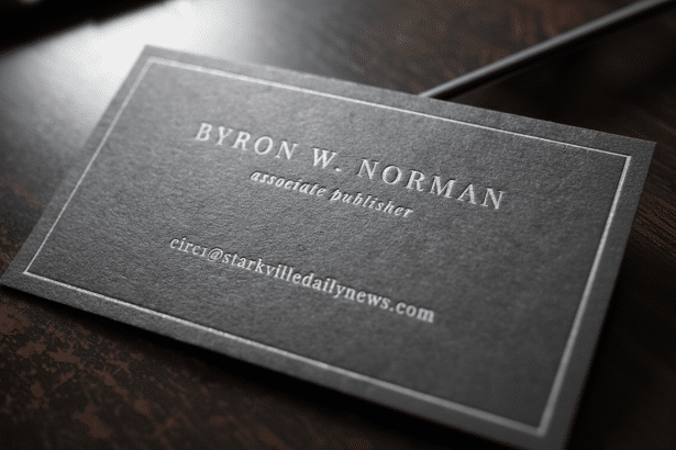 A professional dark gray business card with white embossed text, belonging to Byron W. Norman, associate publisher , with the email cir ci@stark v illedailynews .com, presented on a dark wooden surface. Filename : byron norm anbusiness card. png
