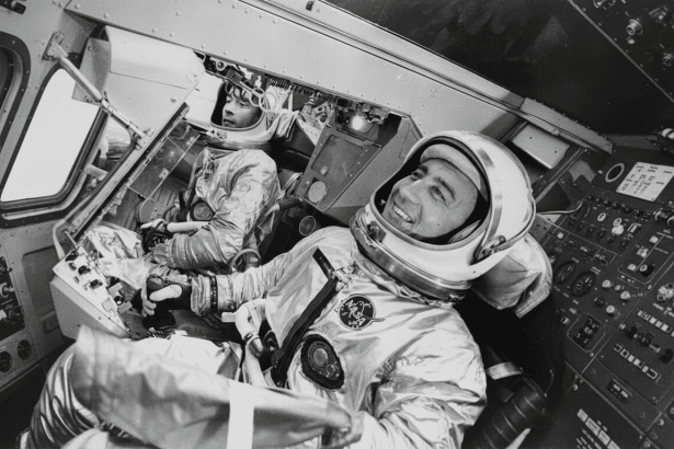 Two astronauts in their spaces uits are seated inside a spacecraft , with one looking forward and the other smiling towards the side .