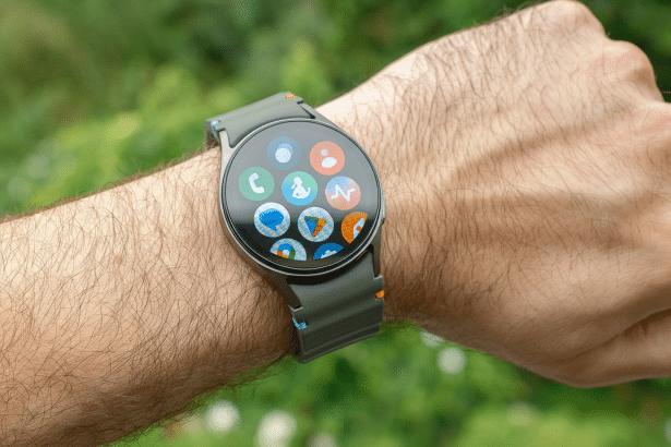 A close-up of a persons arm wearing a smartwatch with various app icons displayed on its screen, set against a blurred green outdoor background.