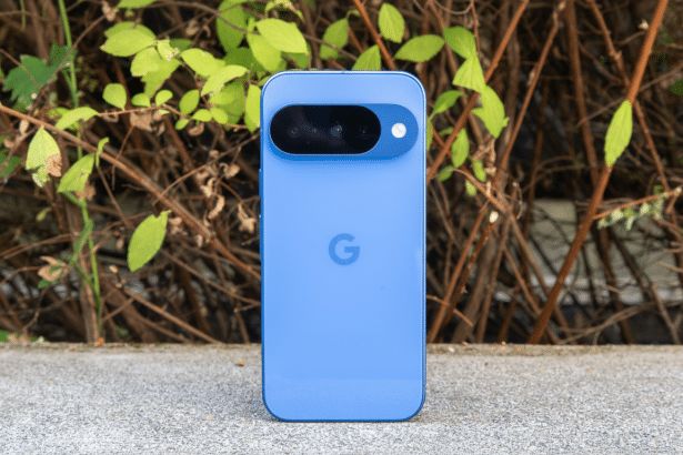 A blue Google Pixel 7 a smartphone is shown upright on a concrete surface with a natural background of green leaves and brown branches. Filename : google pixel7 aprofessional enh ancement.png
