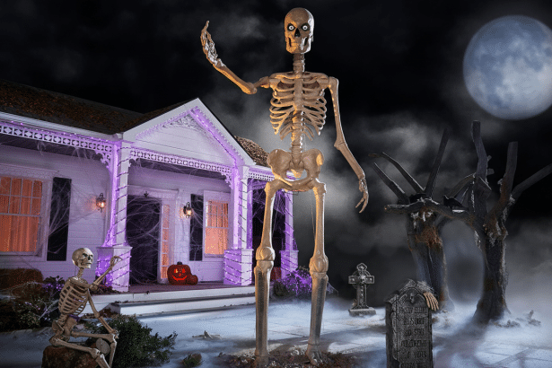 A large skeleton decoration stands in a yard at night, with a smaller skeleton seated nearby . A white house decorated with purple lights, a pumpkin, and spiderwebs is in the background, under a full moon with fog on the ground.