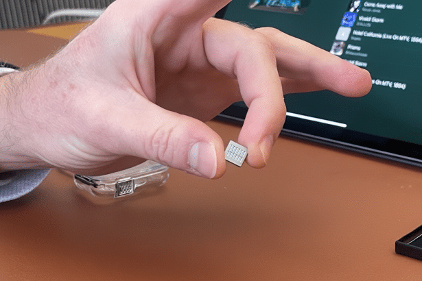 A man 's hand holds a tiny , rectangular electronic component between his thumb and index finger, with a tablet displaying a music playlist in the blurred background. Filename : hand holdingmicro chip.png