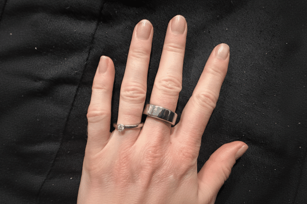 A hand wearing two rings, one a thin silver band with a diamond and the other a wider silver band, against a black fabric background. Filename : hand withrings .png