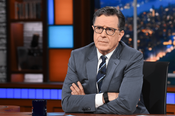 Stephen Colbert sitting at a desk with his arms crossed, wearing glasses and a suit, with a city skyline visible through a window in the background.