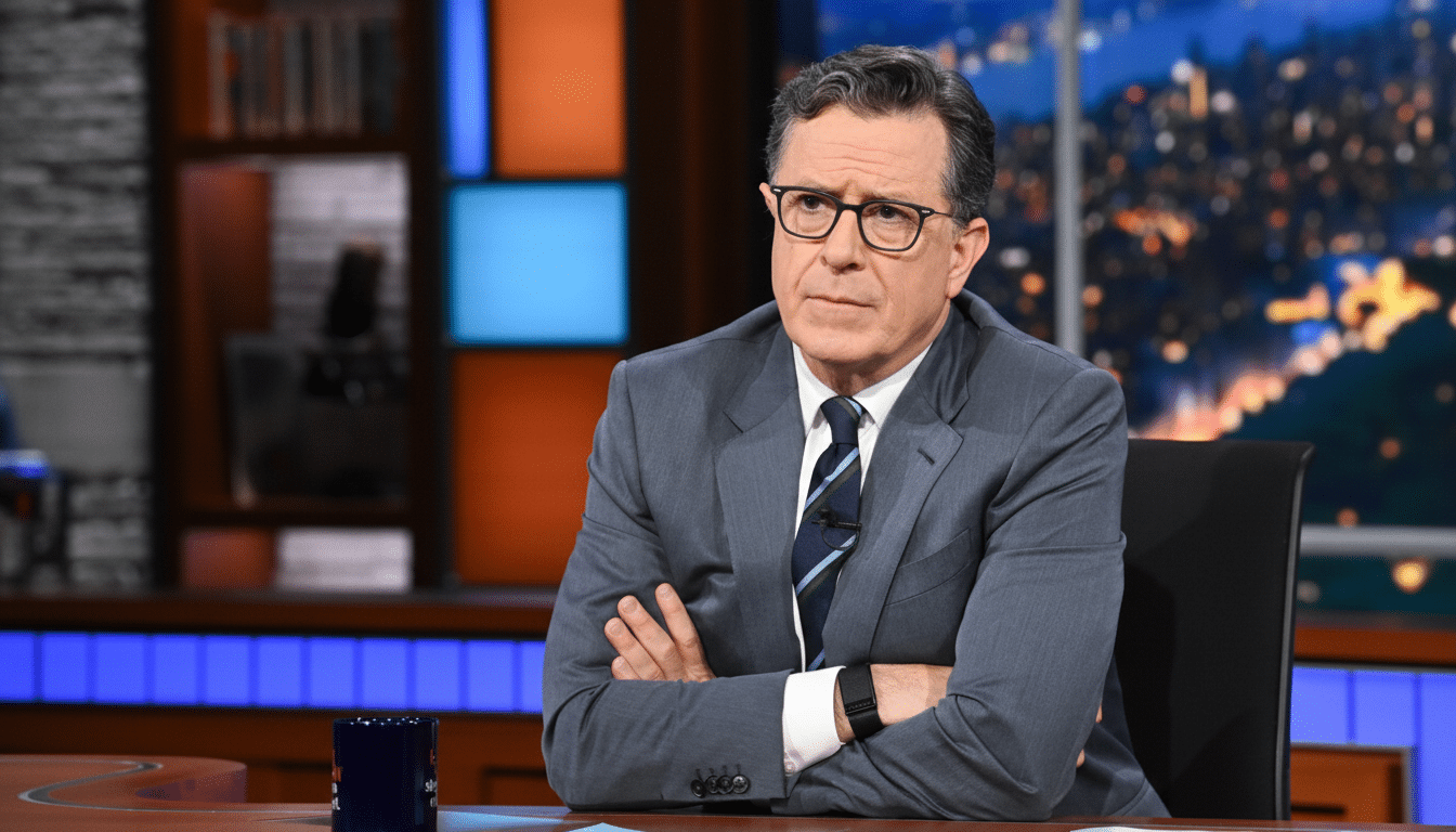 Stephen Colbert sitting at a desk with his arms crossed, wearing glasses and a suit, with a city skyline visible through a window in the background.