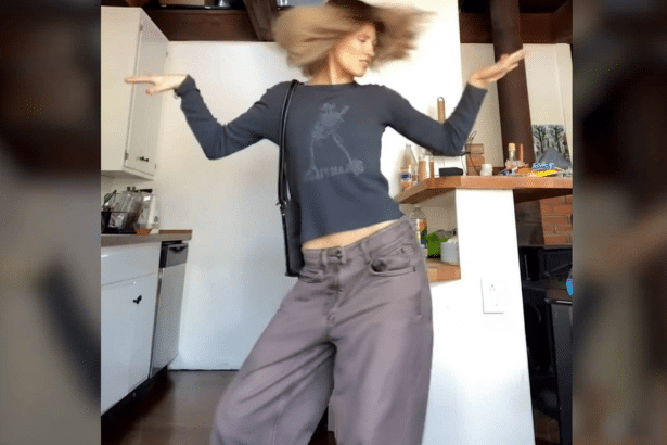 A person with light brown hair dancing in a kitchen , wearing a dark long-sleeved shirt and wide- legged pants.