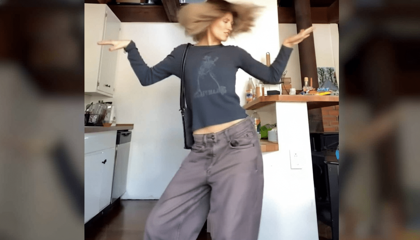 A person with light brown hair dancing in a kitchen , wearing a dark long-sleeved shirt and wide- legged pants.