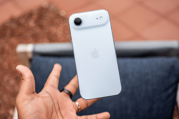 A hand holding a light blue smartphone with an Apple logo on the back, shown outdoors at a 16: 9 aspect ratio.
