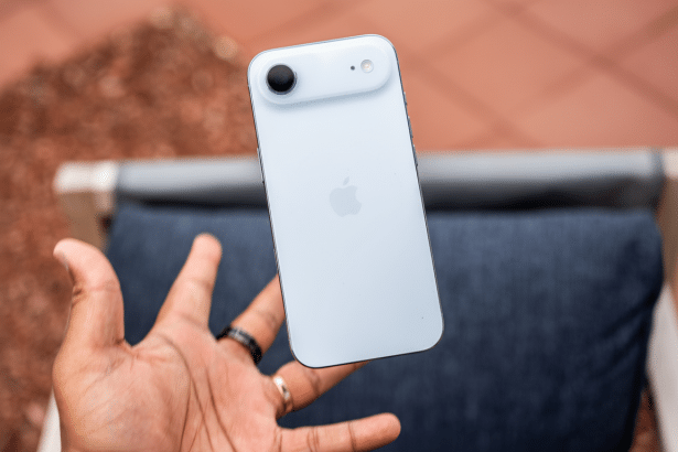 A hand holding a white iPhone 14, with the Apple logo visible on the back, against a blurred outdoor background.