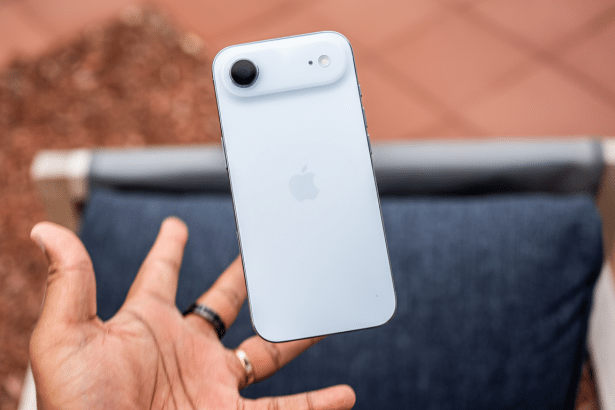 A hand holding a white iPhone 15 vertically , showcasing its back with the Apple logo, camera, and flash , against a blurred background of reddish -brown ground and a blue cushion .