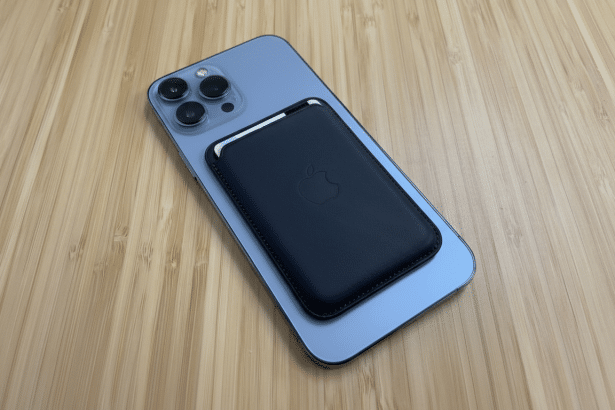 An iPhone 15 Pro Max in blue with a dark blue Apple MagSafe Wallet attached to its back , resting on a wooden surface .
