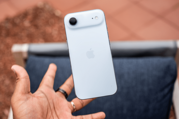 A hand holding a white iPhone 15, with the camera and Apple logo visible on the back, against a blurred outdoor background.