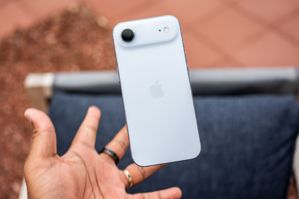 A hand holding a light blue iPhone against a blurred outdoor background of a terracotta- colored surface and a blue cushion .