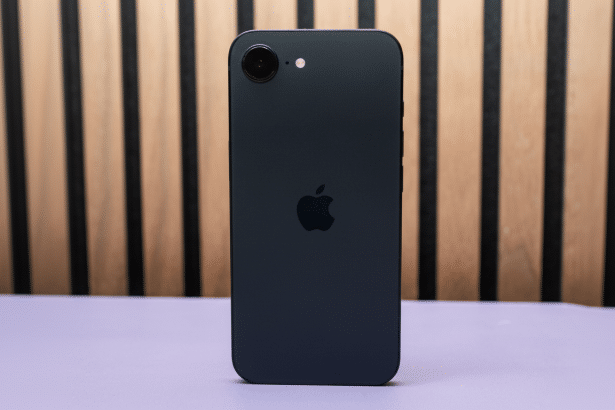 A black iPhone SE ( 3 rd generation) is shown from the back, centered on a light purple surface with a wooden sl atted background. The Apple logo is v