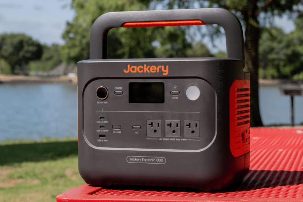 A black and orange Jackery Explorer 10 00 portable power station with multiple outlets and ports, sitting on a red picnic table outdoors with a lake and trees in the background.