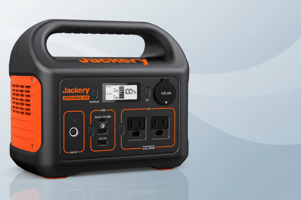 A professional image of a Jackery Explorer 300 portable power station, black with orange accents, against a clean , light grey gradient background.