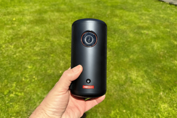 A hand holding a black cylindrical Nebula projector with a red logo against a green grass background. Filename : nebulaproject or16 9. png