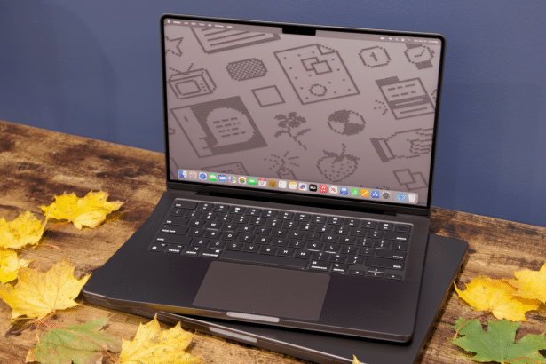 A professional shot of a dark gray MacBook Pro with its screen displaying a patterned wallpaper , resting on a wooden surface adorned with autumn leaves, against a blue wall. Filename : macbook proautumn display .png