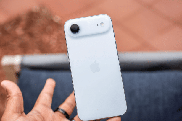 A hand holding a light blue smartphone, showcasing its back camera and the Apple logo, with a blurred outdoor background.
