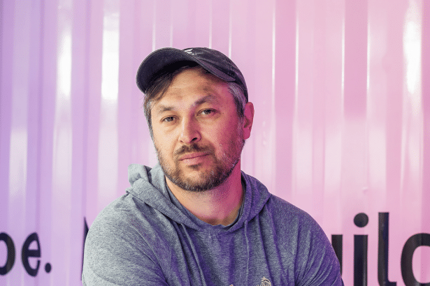 A man with a beard and a baseball cap looks directly at the camera , resized to a 1 6:9 aspect ratio, with the original pink and white striped background maintained .
