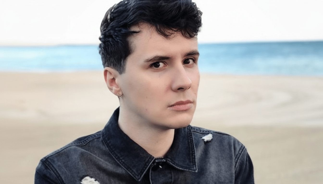 A young man with dark hair and a serious expression , wearing a black denim jacket, stands on a sandy beach with the ocean in the background.