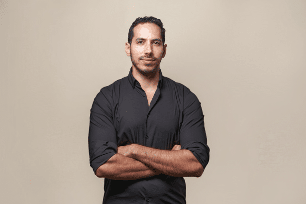 A man with dark hair and a beard, wearing a black shirt, stands with his arms crossed against a plain beige background. Filename : man black shirtarms crossed.png
