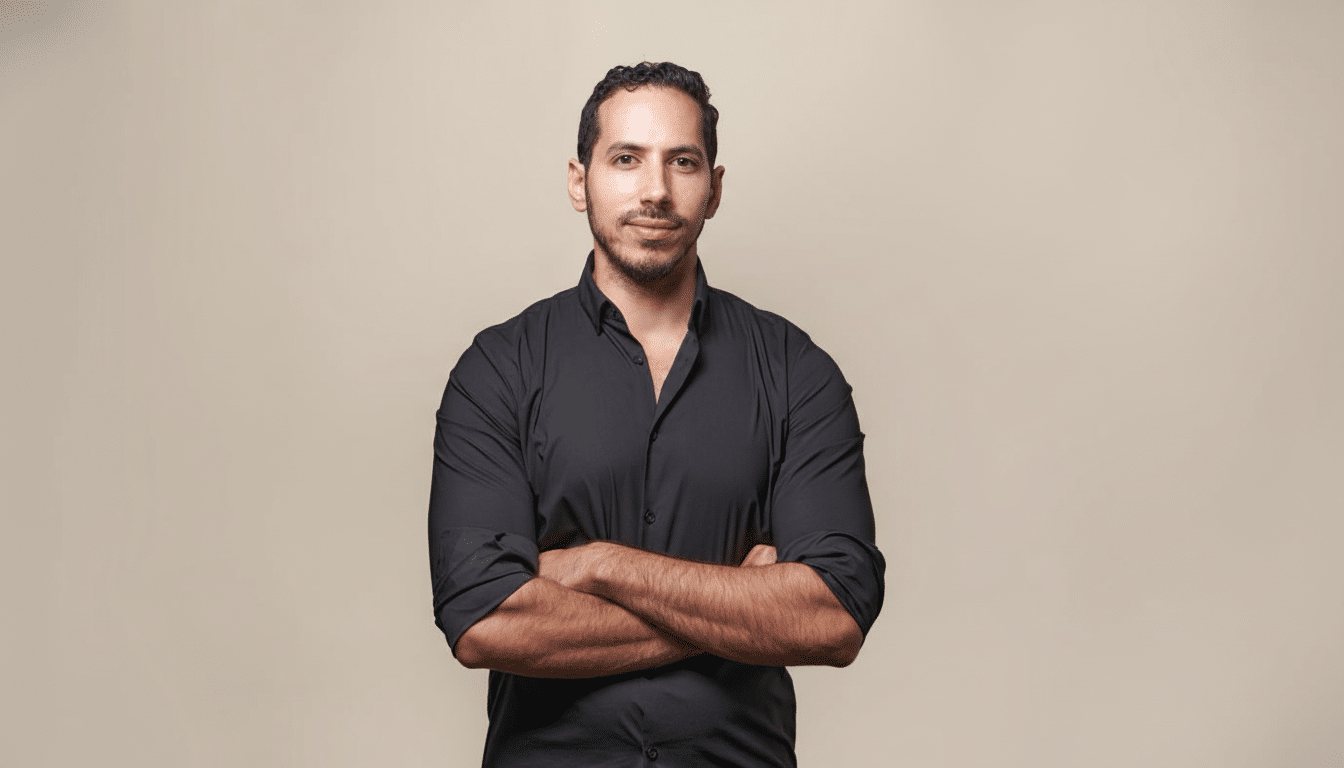 A man with dark hair and a beard, wearing a black shirt, stands with his arms crossed against a plain beige background. Filename : man black shirtarms crossed.png