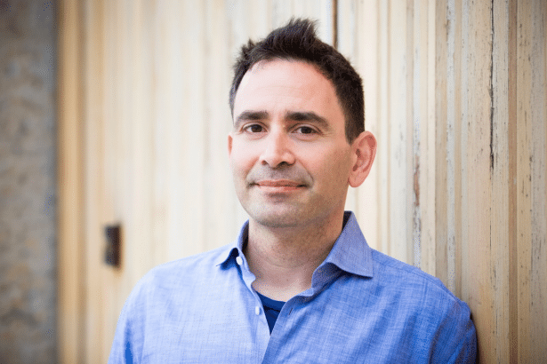 A man in a blue shirt with dark hair smiles , leaning against a light wooden wall.