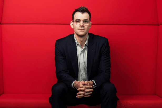 A man in a dark suit and glasses sits against a bright red background , looking directly at the camera with his hands clasped. Filename : man red backgroundprofessional .png