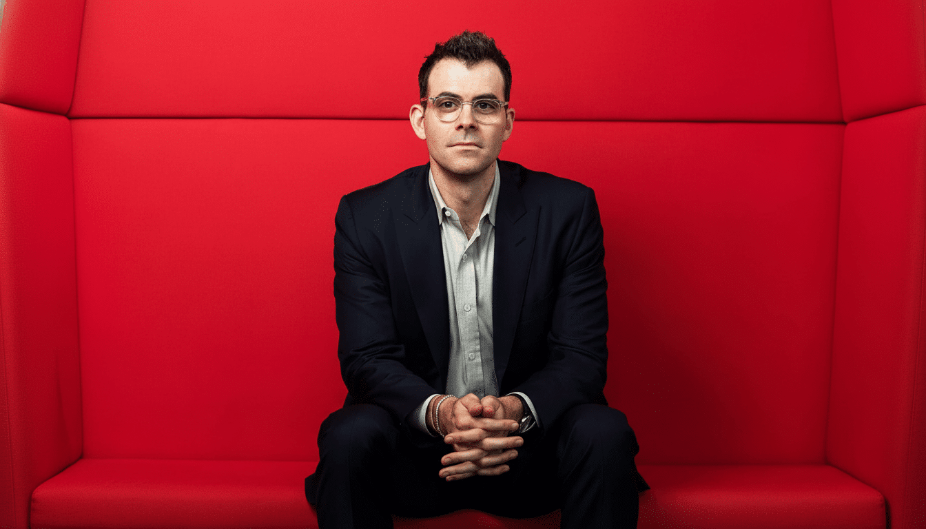 A man in a dark suit and glasses sits against a bright red background , looking directly at the camera with his hands clasped. Filename : man red backgroundprofessional .png