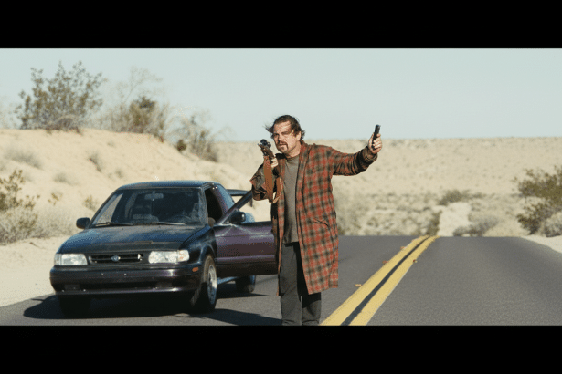 A man in a plaid coat holding a rifle and a phone, standing next to an open car door on a desert road .