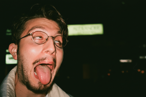 A man with glasses and a beard making a silly face with his tongue out and eyes rolled up , against a dark , out -of-focus background. Filename : man silly facedark background.png