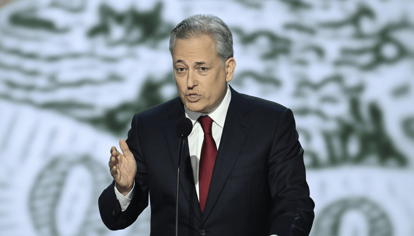 A man in a dark suit and red tie speaking at a podium with a microphone, gesturing with his left hand.